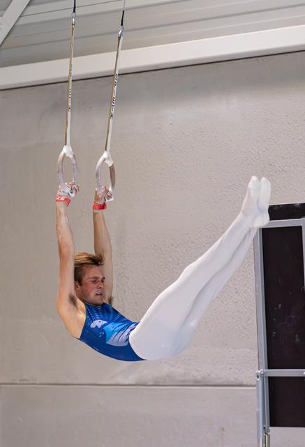 Young man executes a horizontal hold on still rings, wearing blue leotard and white pants in indoor training facility