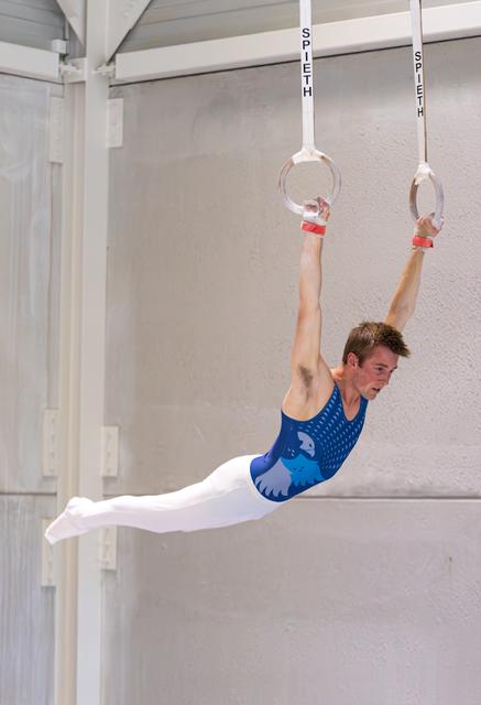 Male gymnast in blue leotard performs horizontal position on still rings, demonstrating exceptional strength and form