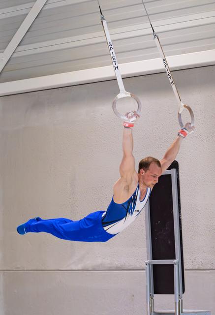 Male gymnast performing horizontal hold on rings, body extended parallel to ground in blue and white uniform