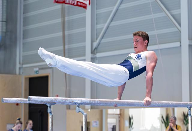 Male gymnast performing a V-sit hold on parallel bars, demonstrating strength and control in an indoor training facility