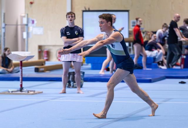 Male gymnast performs floor exercise with arms extended forward, focused expression, training facility with teammates in background