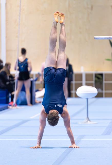Male gymnast performs a vertical handstand on floor exercise mat during indoor competition, legs extended straight upward