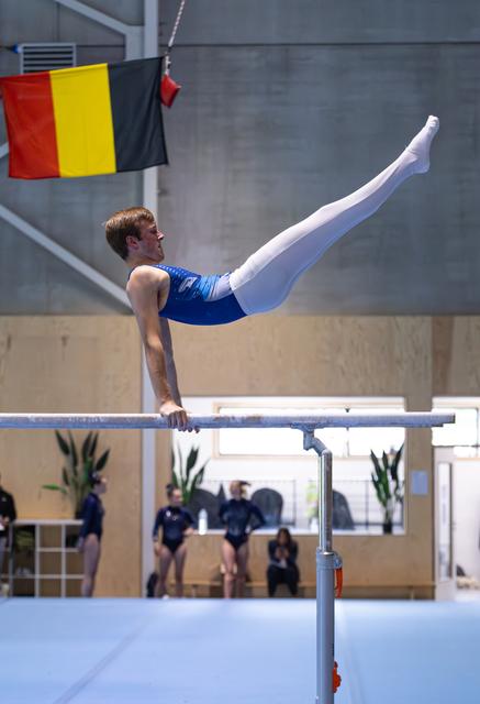 Male gymnast performing horizontal bar routine with elevated body position, Belgian flag visible above in training facility