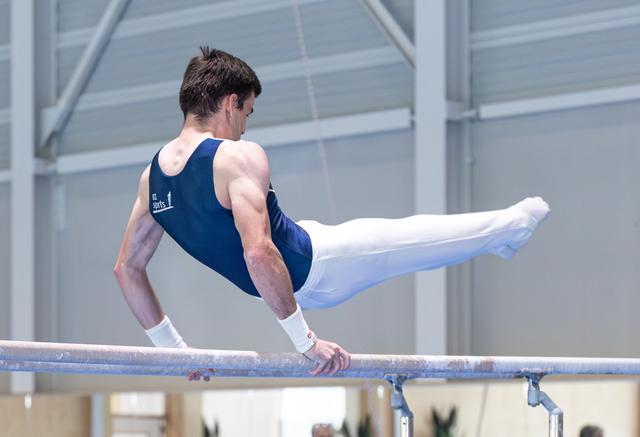 Male gymnast performing horizontal bar routine with legs extended, displaying strength and control in indoor training facility