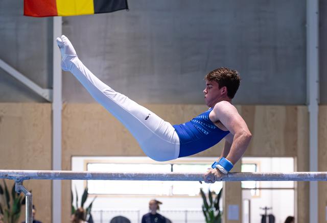 Male gymnast performing a pike hold on horizontal bar, wearing blue uniform, Belgian flag visible above