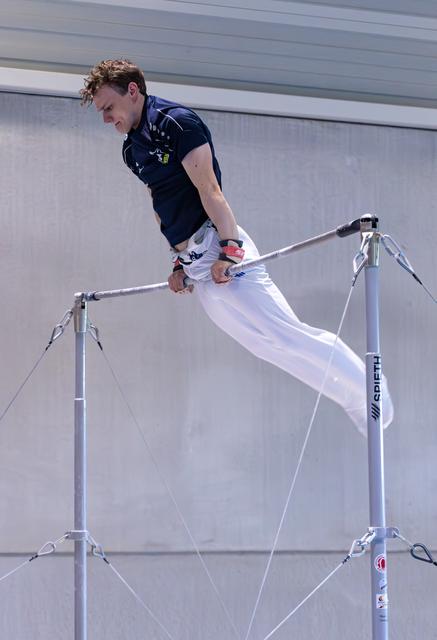 Male athlete performs on horizontal bar, body extended horizontally in white pants and navy shirt against industrial wall
