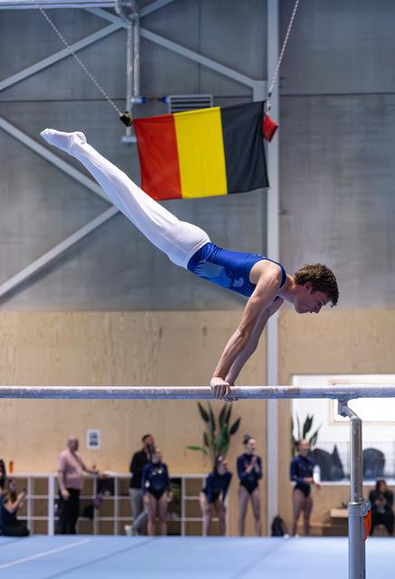 Male gymnast performs a horizontal hold on parallel bars with Belgian flag hanging in the background at indoor venue