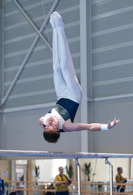 Male gymnast performs inverted flip above horizontal bar, arms extended, in indoor training facility
