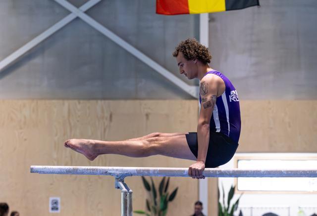 Male athlete performing L-sit on parallel bars, displaying strength and control in purple sleeveless uniform