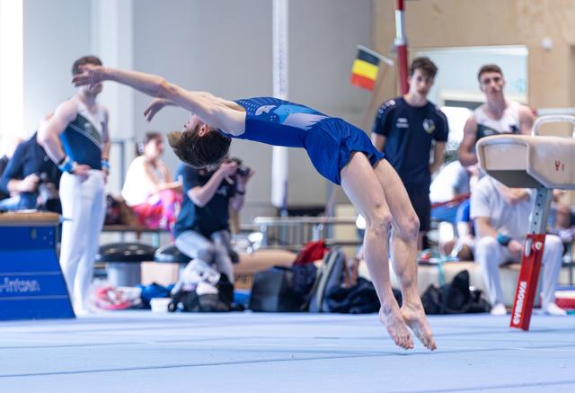 Male gymnast performs backward somersault on floor exercise mat during indoor training event with teammates watching