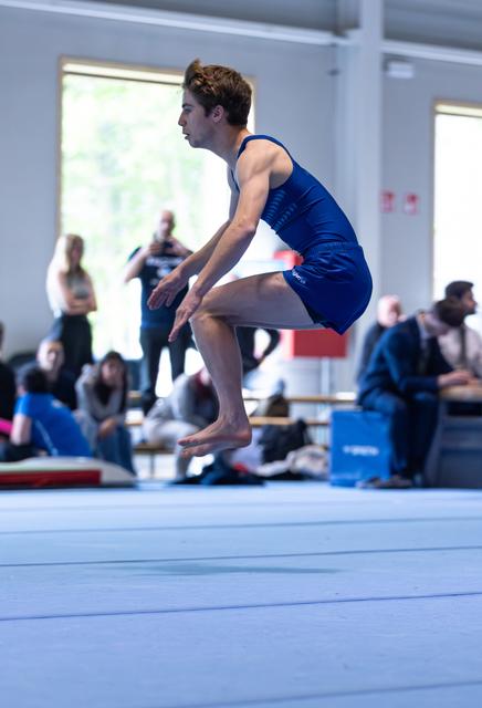 Male gymnast performing a jump during floor routine, body in tucked position, spectators watching in background