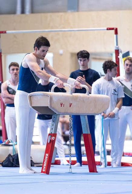 Athlete performs pommel horse routine with focused concentration while teammates watch in background at training facility