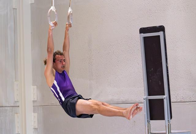 Male gymnast performs L-sit hold on rings wearing purple singlet during indoor training session