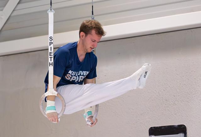 Male gymnast performing on rings in white pants and blue team shirt, demonstrating strength hold during indoor training