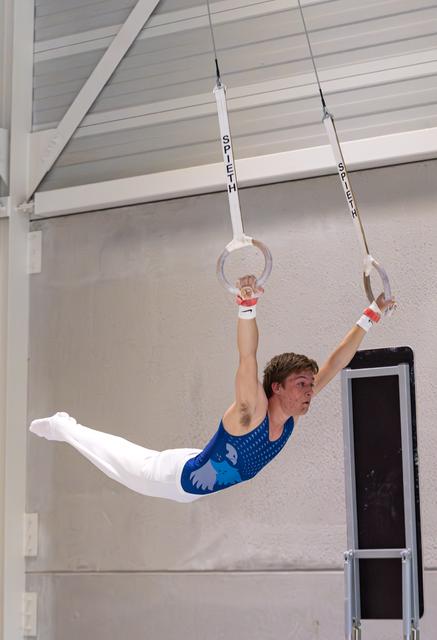 Male gymnast performing horizontal hold on rings, wearing blue leotard and white pants in training facility