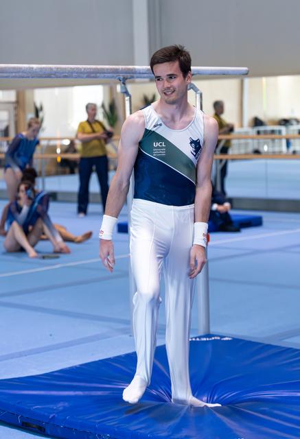 Gymnast in UCL uniform stands confidently on blue mat, smiling after completing his routine before audience