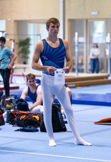 Young male gymnast in blue leotard stands confidently with competition number 11 pinned to his white pants in training facility