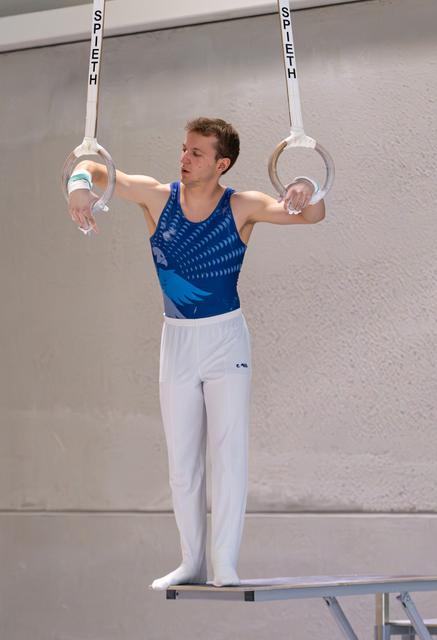 Young male athlete performing on still rings, arms extended, wearing blue patterned leotard and white pants