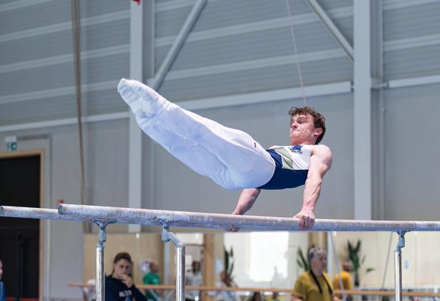 Male gymnast performs horizontal bar routine with extended body position, displaying strength and control during indoor event