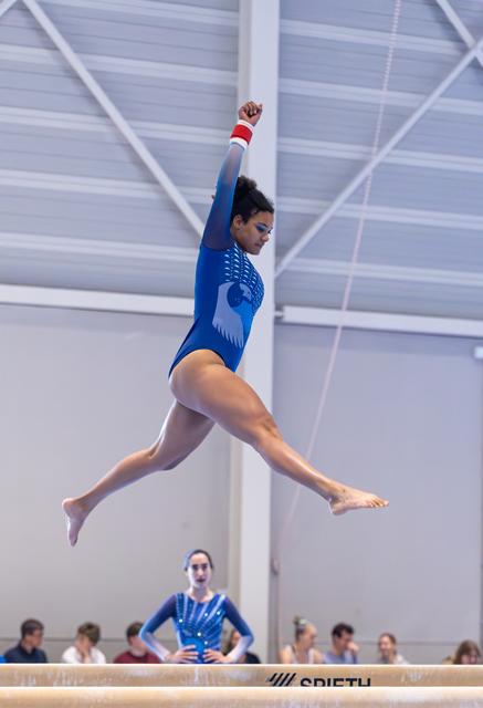 Gymnast performing an impressive mid-air leap above balance beam, showcasing powerful form and extension during routine