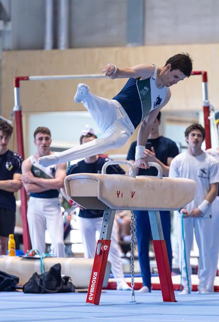 Athlete performing spread-legged straddle on pommel horse, body horizontal in air, with spectators watching intently