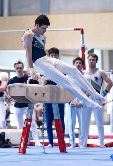 Athlete performs pommel horse routine with legs extended horizontally while teammates watch intently in background
