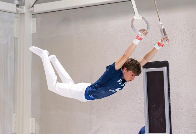 Young athlete demonstrates impressive horizontal hold on still rings, arms extended overhead, wearing blue uniform and white pants