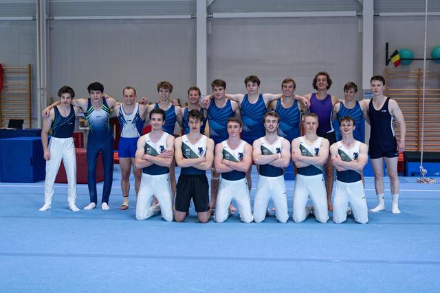 Two rows of young male gymnasts pose together on blue floor mat in training facility, wearing various leotards