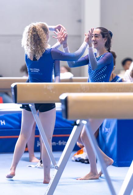 Two gymnasts in blue leotards share an enthusiastic high-five near the balance beam, celebrating together with bright smiles