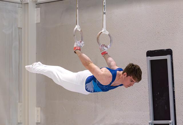 Young male gymnast performing horizontal hold on still rings, demonstrating strength and control in white outfit