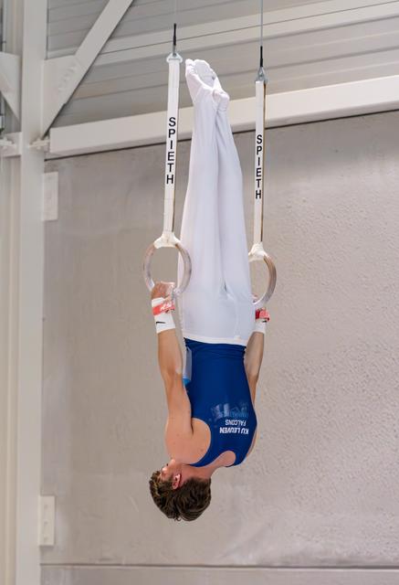 Young gymnast performs an inverted hang on still rings, body fully extended upside down in white and blue attire