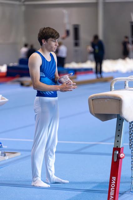 Young gymnast in blue leotard stands focused beside pommel horse, hands clasped in preparation before his routine