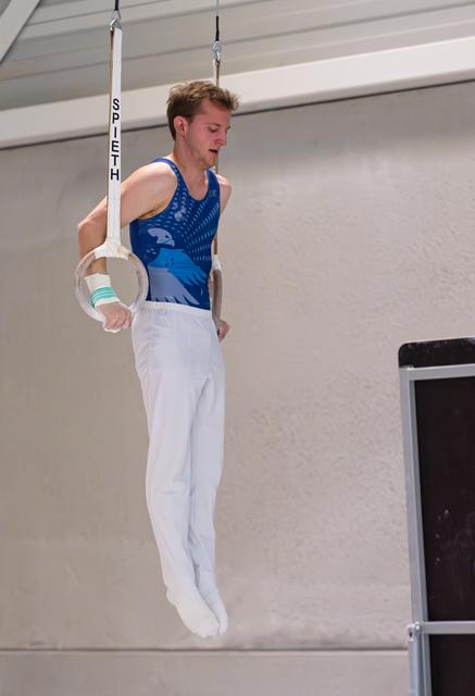 Male gymnast performing on still rings in blue leotard, focused expression while suspended in air at indoor venue