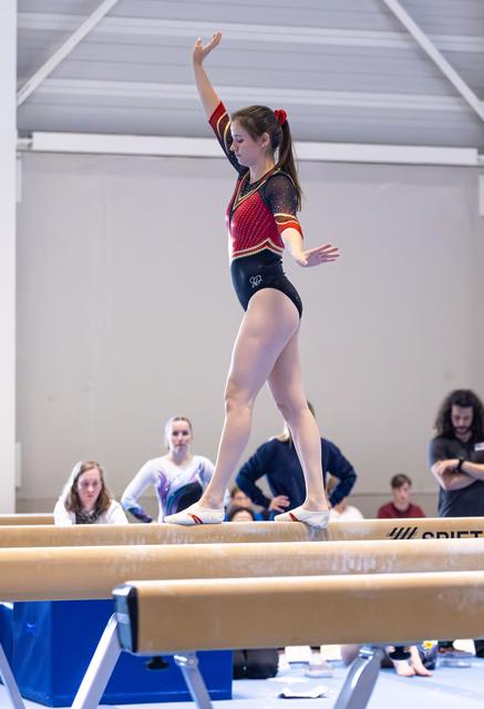 Gymnast performs on balance beam with graceful arm position, wearing red and black leotard, while teammates watch below