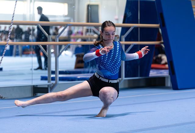 Young gymnast performs a split leap during floor routine, wearing blue leotard with focused expression in training facility
