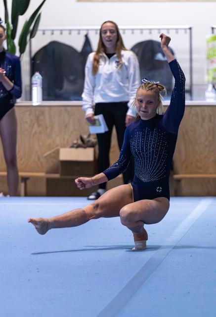 Young gymnast performing a floor routine landing pose in navy leotard while coach observes in background