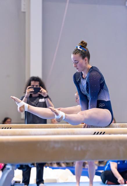 Young gymnast in blue leotard performs splits position on parallel bars while spectator photographs in background