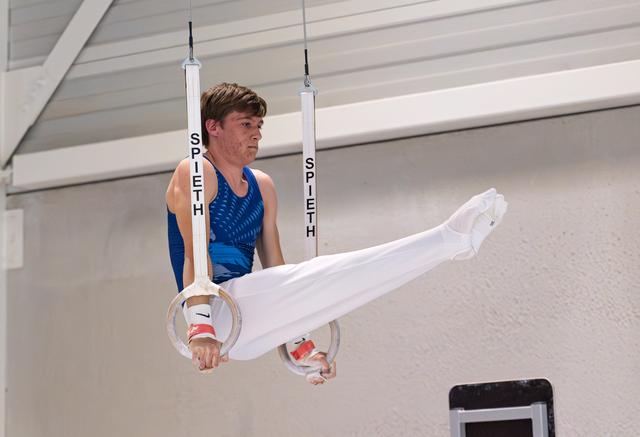 Male gymnast in blue leotard performing on rings with extended legs in white pants, focused expression in training facility