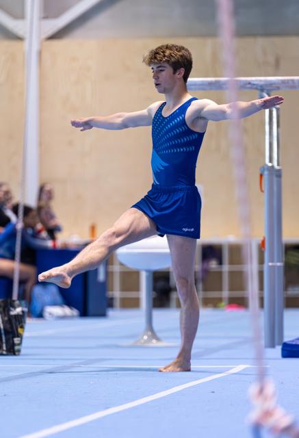 Male gymnast performing floor routine with arms extended, executing arabesque pose on blue mat in indoor arena