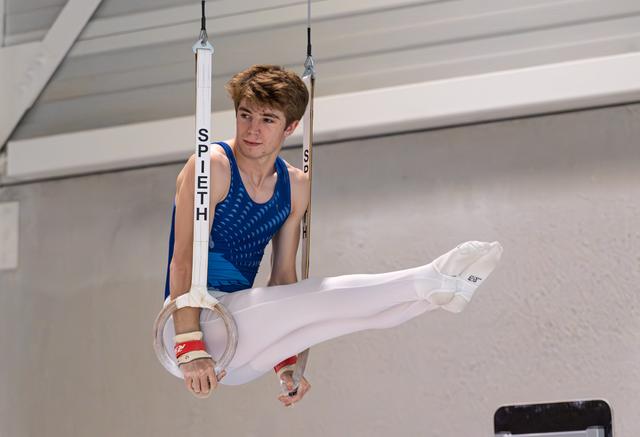 Male gymnast in blue leotard executes a straight-leg position on rings, displaying focus and strength during his routine.