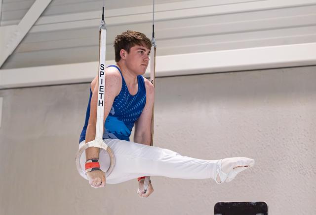 Young male gymnast performing a horizontal hold on still rings, wearing a blue leotard and white pants, looking focused