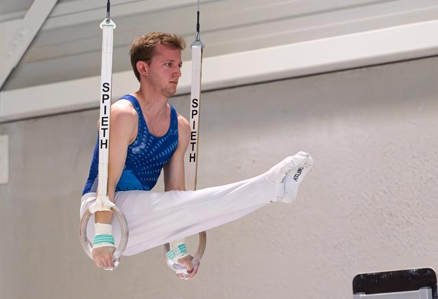 Young male gymnast performing a horizontal leg position on still rings, wearing blue leotard and white pants